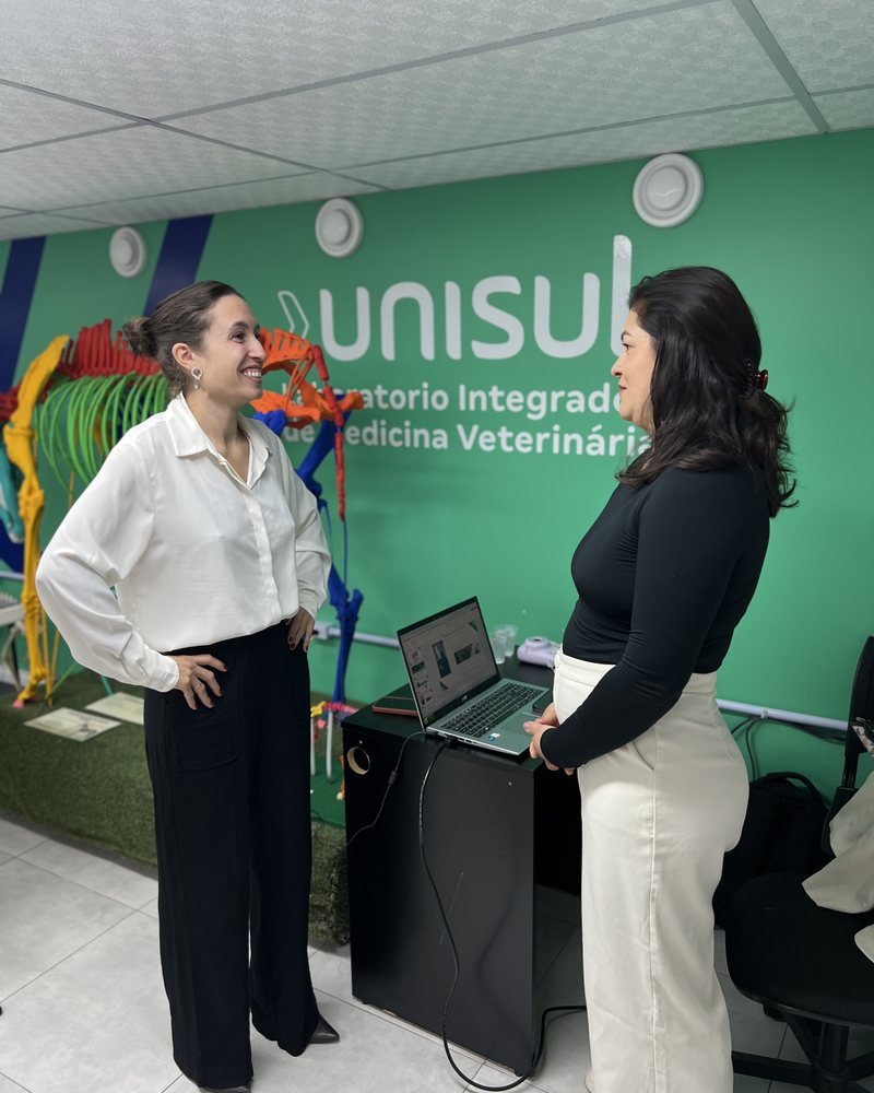 Duas pessoas em pé, voltadas uma para a outra, junto a um notebook sobre um suporte.