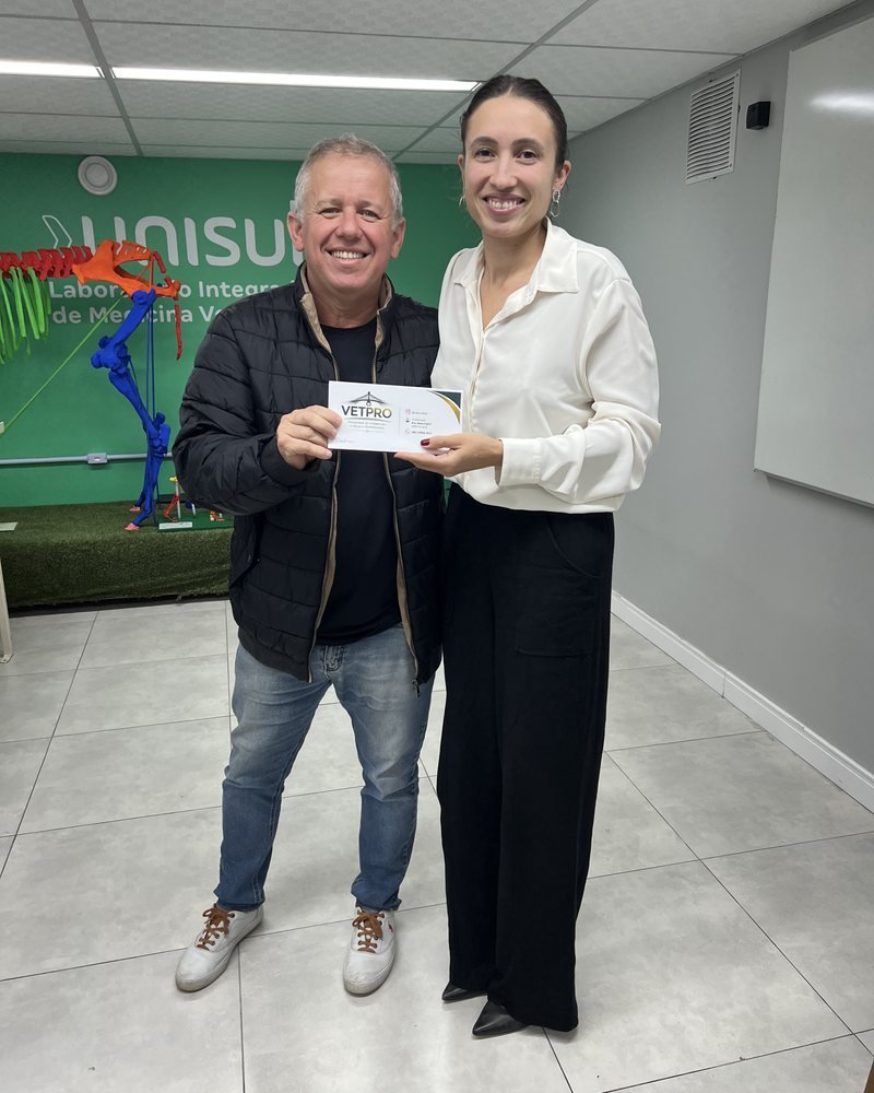 Duas pessoas voltadas para a câmera; uma delas segura um cartão impresso.