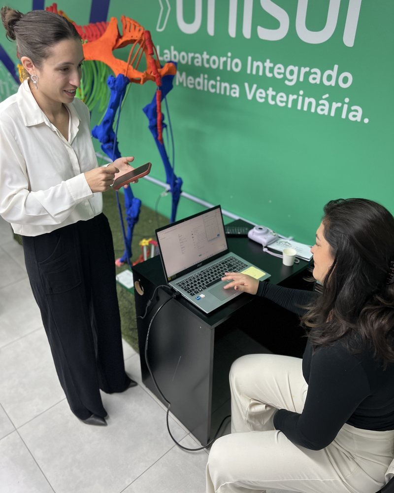 Uma pessoa em pé junto a outra sentada diante de um notebook; ao fundo, banner do laboratório de medicina veterinária.